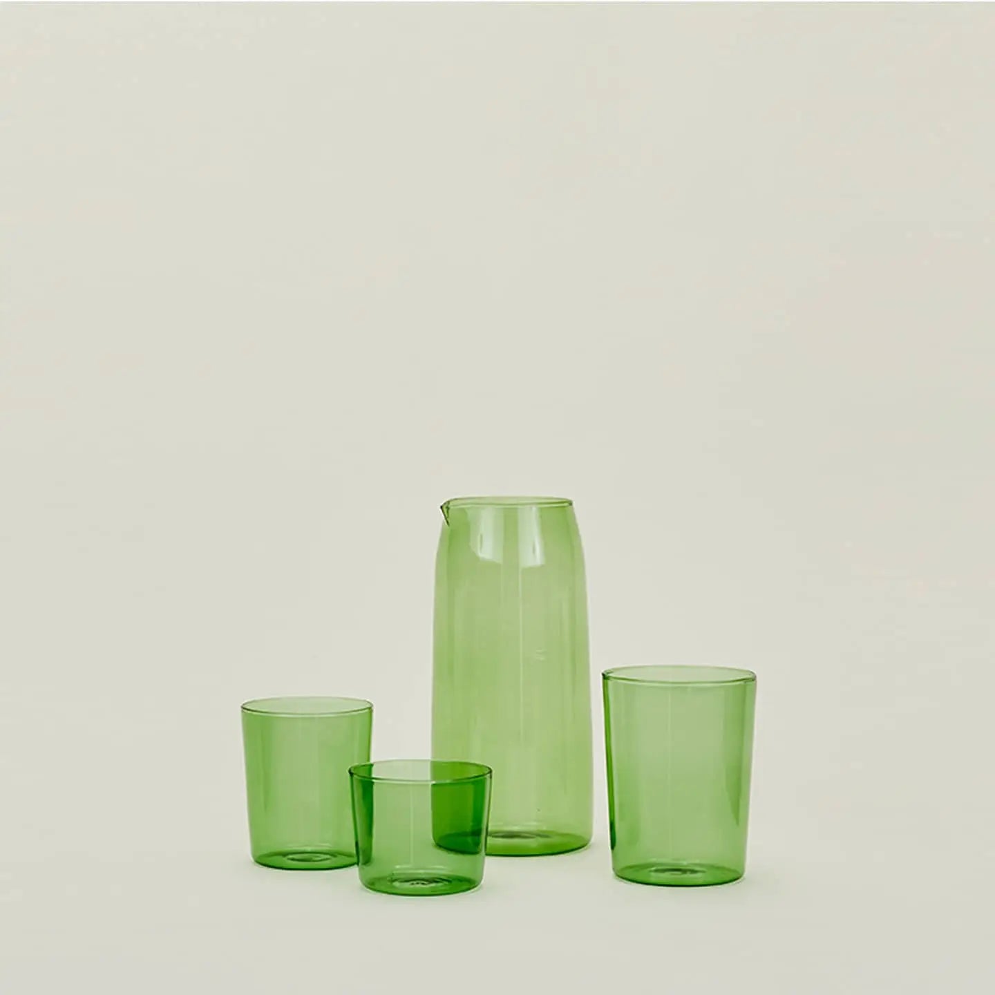 A set of four small translucent green Hawkins New York Essential Glassware glasses are arranged in front of a matching pitcher, all displayed against a plain light background.