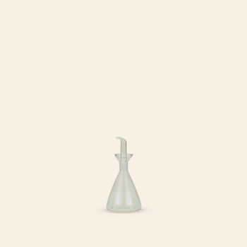 The Hawkins New York Essential Kitchen Bottle, Medium, is a clear, modern bottle with a narrow neck, funnel-shaped top, and precision drizzle spout. It sits centered on a plain light beige background for a minimalistic look.