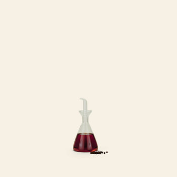 The Hawkins New York Essential Kitchen Bottle, Medium, with modern styling and a precision drizzle spout, holds red liquid—possibly vinegar—and sits on a light surface beside peppercorns against a pale background.