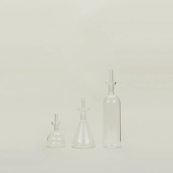 A Hawkins New York Essential Kitchen Bottle (Medium) stands against a light background, showcasing its transparent design, modern styling, and precision drizzle spout with stopper.