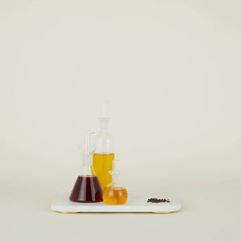 Three Hawkins New York Essential Kitchen Bottles, Medium, filled with dark brown, yellow, and amber liquids, sit on a white tray. A pile of black peppercorns is beside them, all set against a plain light background.