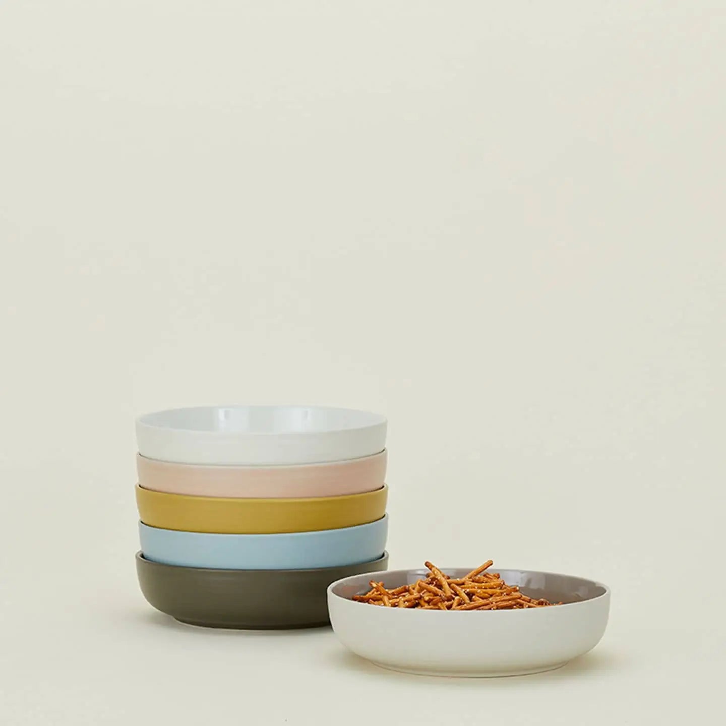 A set of four Hawkins New York Essential Low Bowls in soft hues sits beside a gray bowl with crispy snack sticks, creating a stylish dining scene against a plain beige background.
