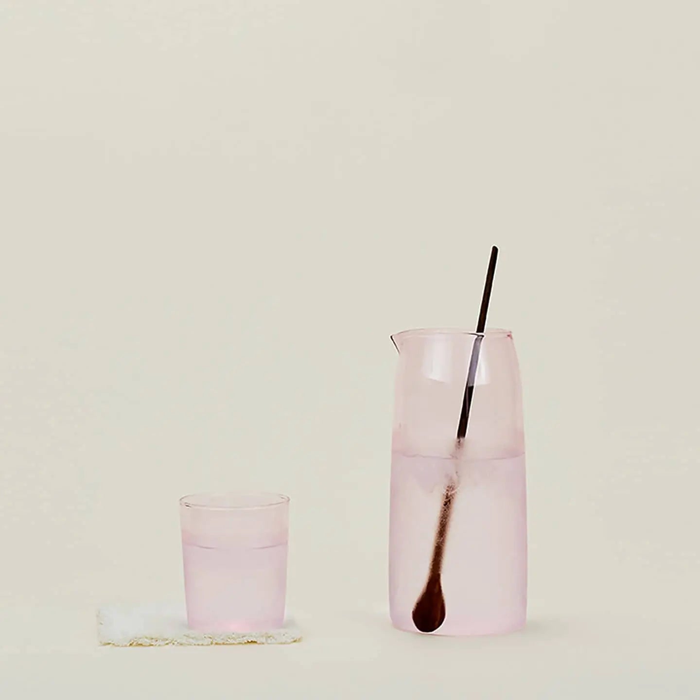 The Hawkins New York Essential Pitcher, filled with a light pink drink and a long spoon, sits beside its matching glass on a soft coaster, all set against a simple beige background.
