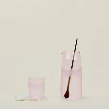 The Hawkins New York Essential Pitcher, filled with a light pink drink and a long spoon, sits beside its matching glass on a soft coaster, all set against a simple beige background.