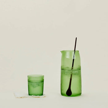 A Hawkins New York Essential Pitcher filled with ice water and a long black stirring spoon sits beside a green glass tumbler with ice on a folded white napkin, all set against a plain light background.