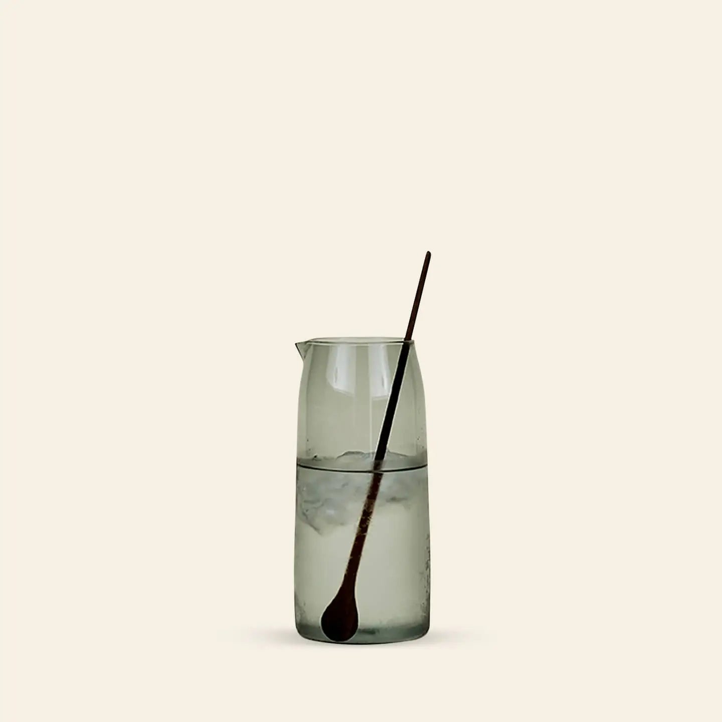 The Hawkins New York Essential Pitcher by Hawkins New York, made of clear glass, is filled with ice water and sits on a light surface against a plain beige background, with a long dark mixing spoon inside.