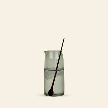 The Hawkins New York Essential Pitcher by Hawkins New York, made of clear glass, is filled with ice water and sits on a light surface against a plain beige background, with a long dark mixing spoon inside.