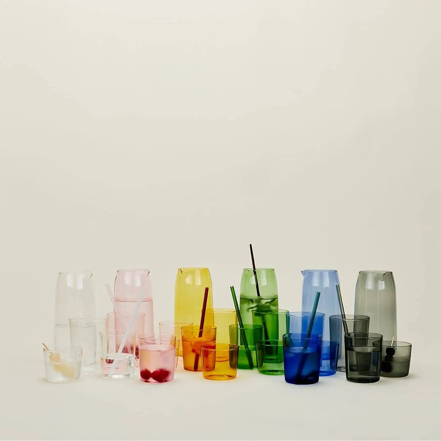 The Hawkins New York Essential Pitchers, each with matching glass straws, are arranged by color—clear, pink, yellow, green, blue, and gray—set against a plain beige background.