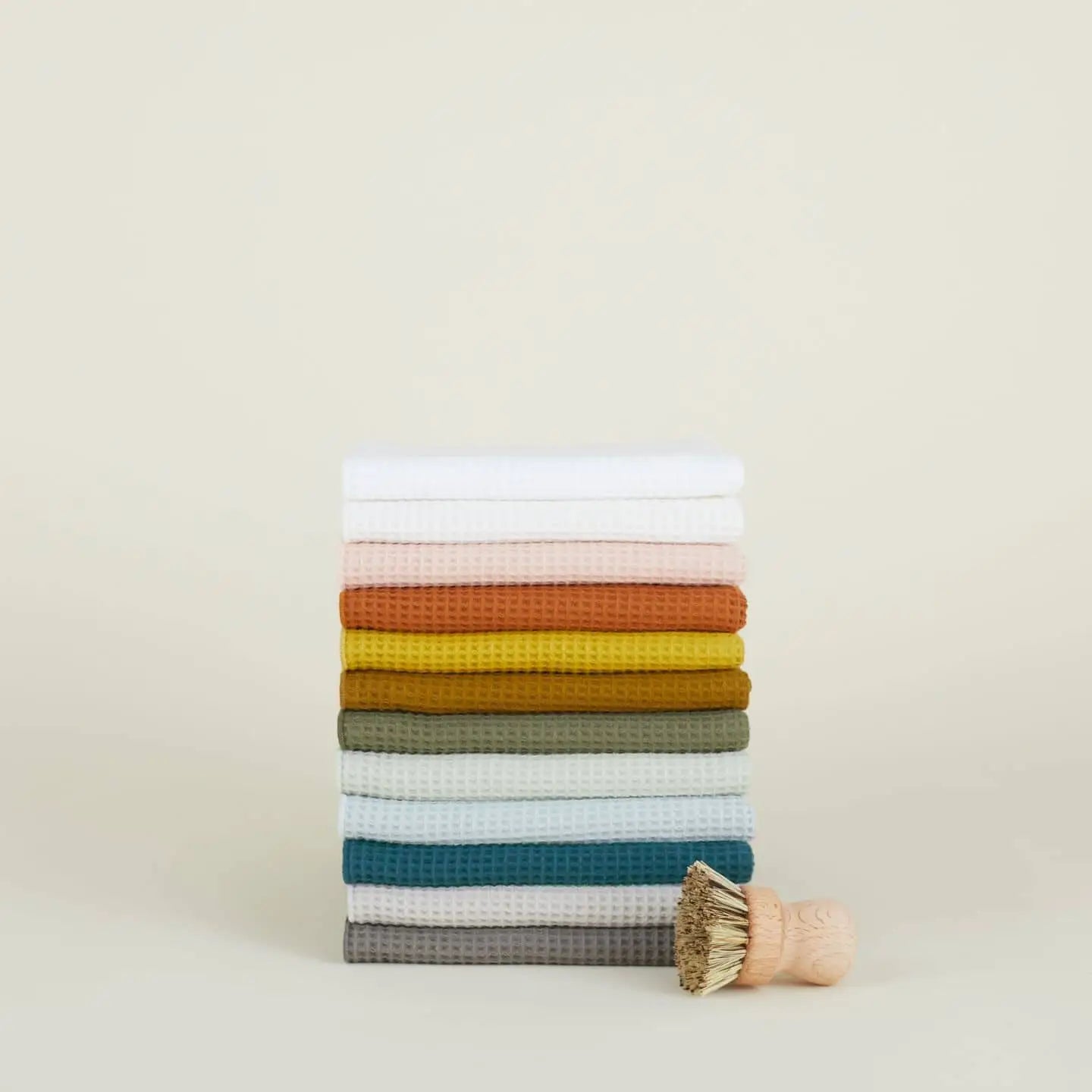 A set of Hawkins New York Essential Waffle Dish Towels in assorted colors is stacked upright, with a small wooden scrub brush in front, all set against a plain, light background.