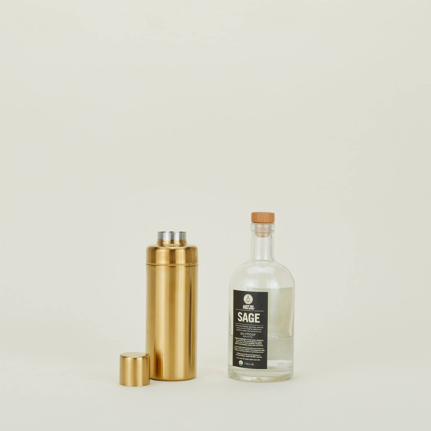 A Hawkins New York Simple Cocktail Shaker with its lid sits beside a clear SAGE bottle with a wooden cap, both set on a plain, light background at The Stacked Store.