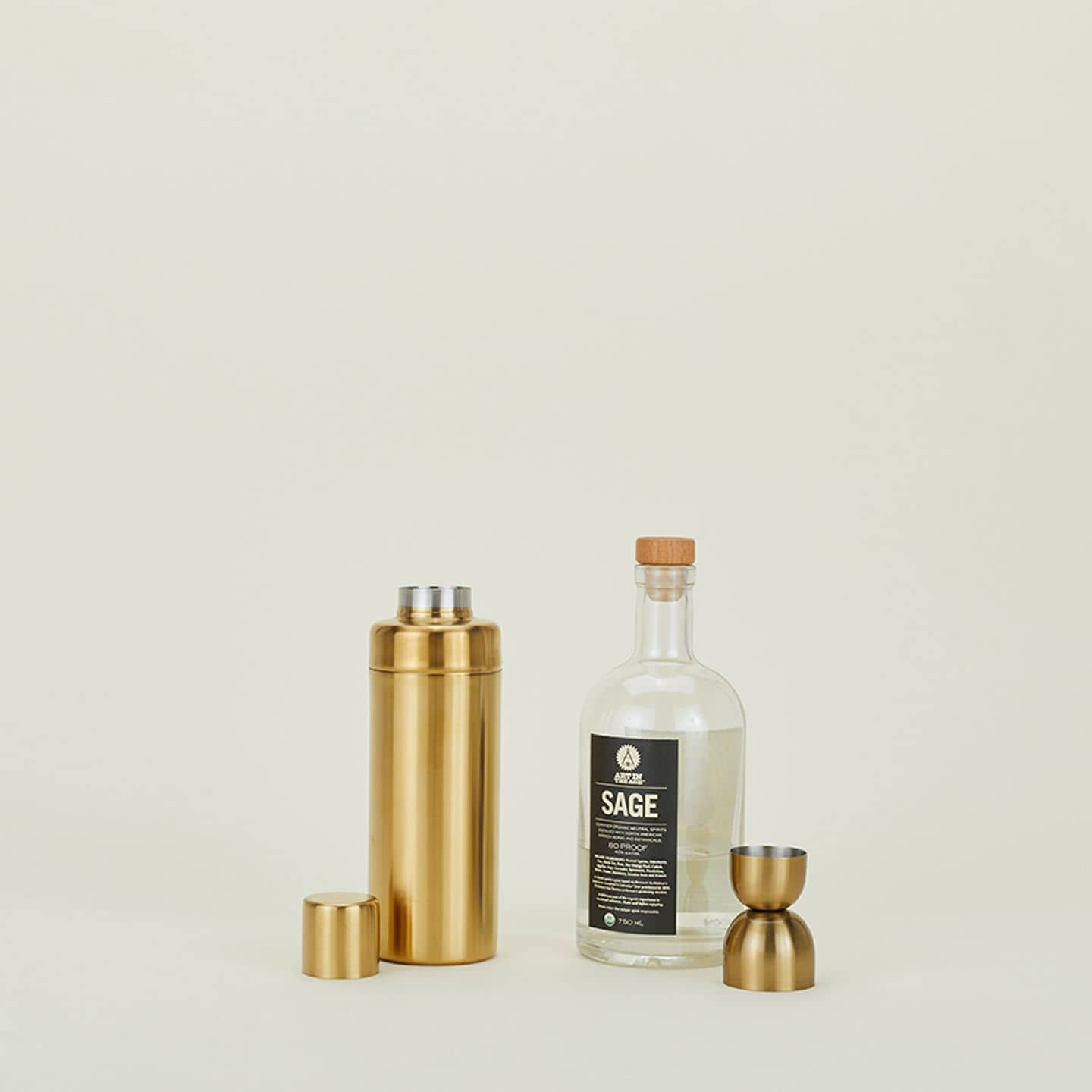 A Hawkins New York Simple Cocktail Shaker with its cap off stands next to a clear glass bottle labeled SAGE and a gold double-sided jigger, all set against a plain light background.