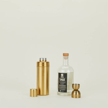 A Hawkins New York Simple Cocktail Shaker with its cap off stands next to a clear glass bottle labeled SAGE and a gold double-sided jigger, all set against a plain light background.
