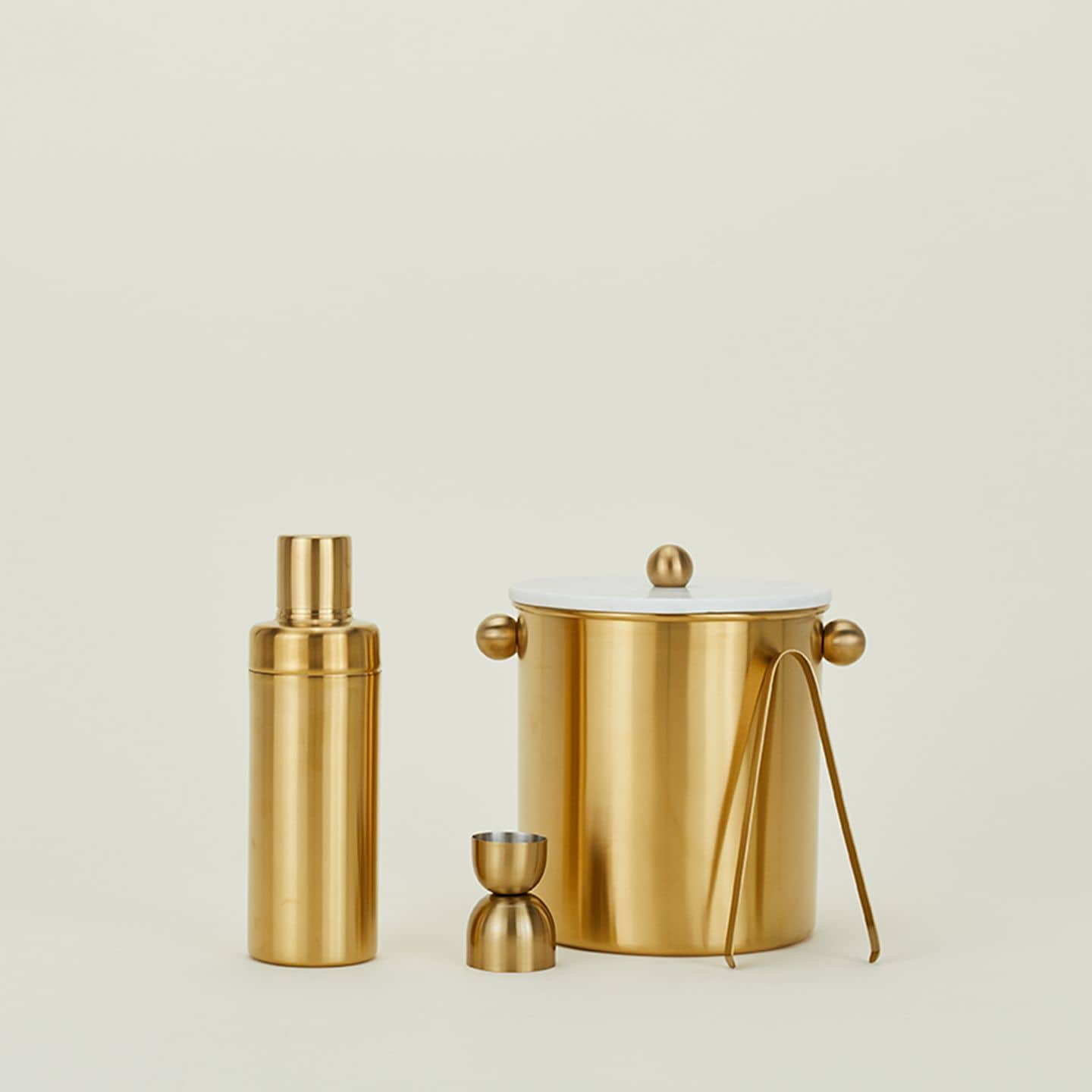 A gold-toned bar set from The Stacked Store features the Hawkins New York Simple Cocktail Shaker, double-sided jigger, ice bucket with tongs, and white lid—beautifully displayed against a plain light background.