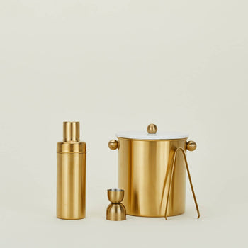 A gold-toned bar set from The Stacked Store features the Hawkins New York Simple Cocktail Shaker, double-sided jigger, ice bucket with tongs, and white lid—beautifully displayed against a plain light background.