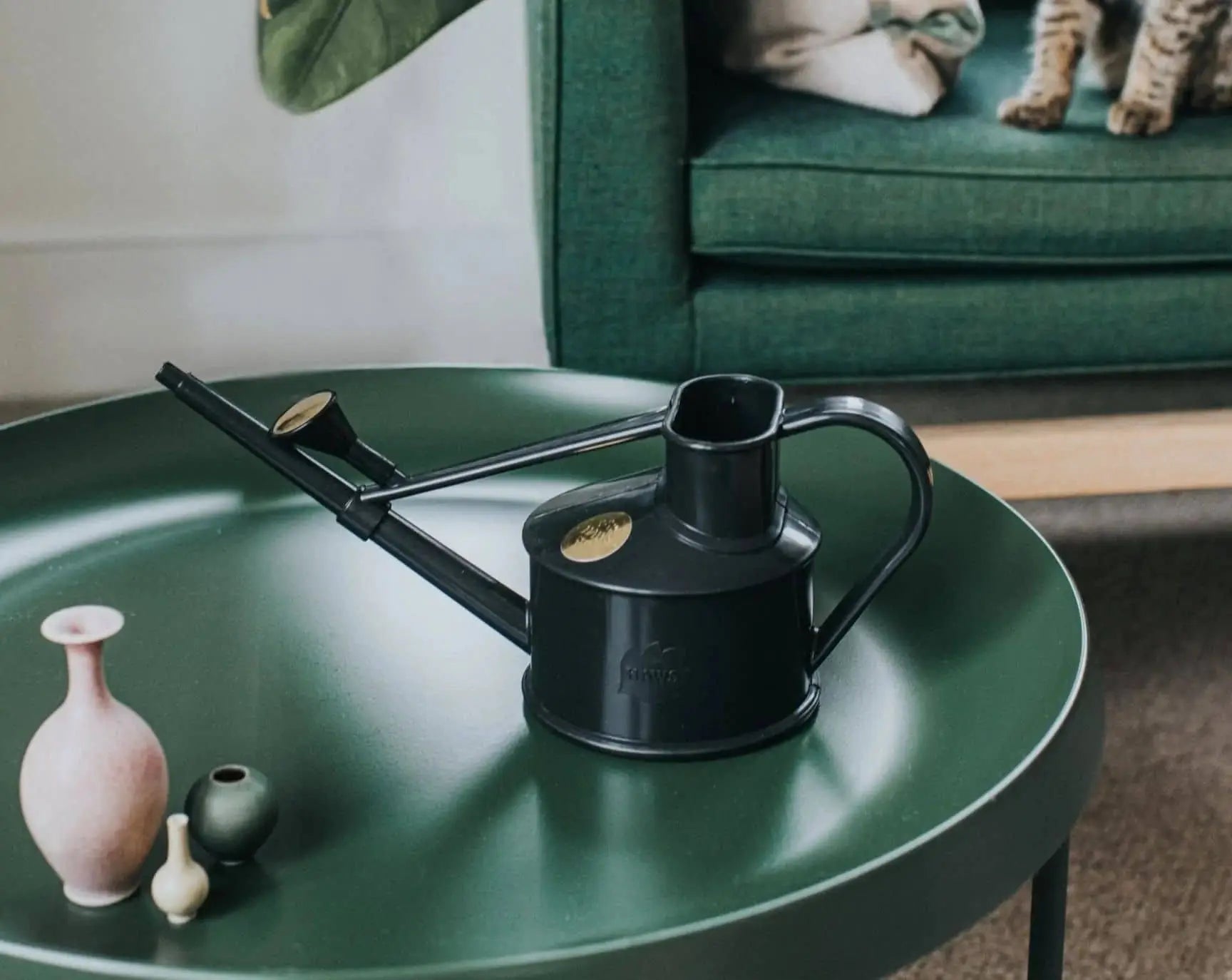 The Haws The Langley Sprinkler, a black watering can with an adjustable head, rests on a round dark green table beside three small decorative vases. Behind them, a green sofa with a cat’s paws shows above the brown carpet.