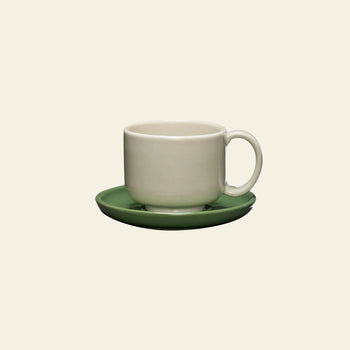 The Hübsch Amare Cup With Saucer by Hübsch, featuring a plain white ceramic cup on a green saucer, sits against a light beige background. This retro-inspired set is simple, unadorned, and dishwasher safe.