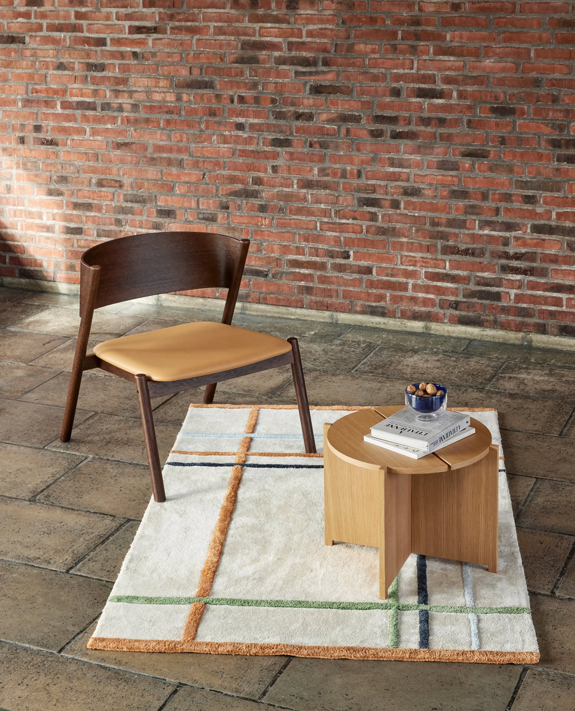 A modern wooden chair with a tan cushion sits on a patterned rug beside the Hübsch Dash Side Table by Hübsch, which is topped with books and a small bowl, all set against a red brick wall for minimalist style.
