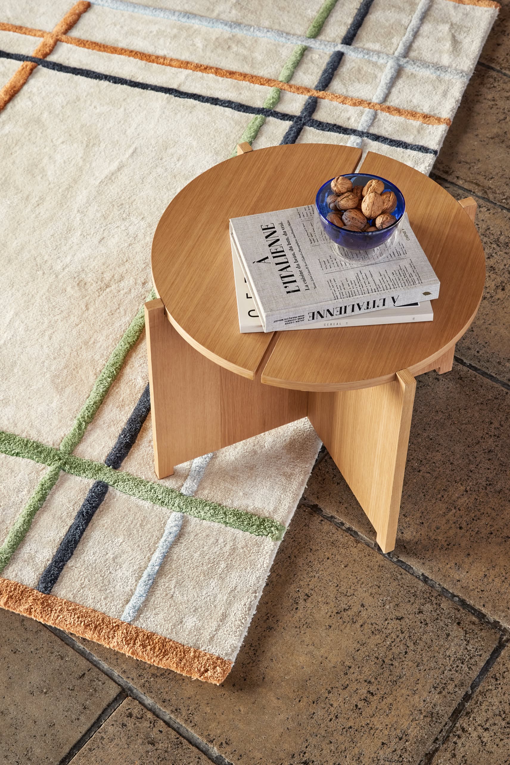 A round Hübsch Dash Side Table by Hübsch, made from FSC-certified wood, stands on a tiled floor and a cream rug with green, orange, and blue grid lines. The table holds a blue bowl of walnuts and two stacked books.