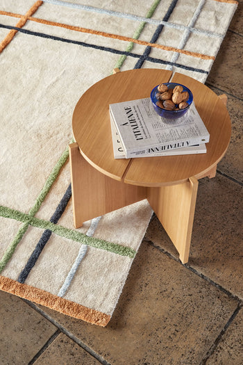 A round Hübsch Dash Side Table by Hübsch, made from FSC-certified wood, stands on a tiled floor and a cream rug with green, orange, and blue grid lines. The table holds a blue bowl of walnuts and two stacked books.