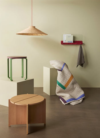Minimalist room with a wooden stool, Hübsch Dash Side Table by Hübsch (green legs, FSC-certified wood), large woven lamp, folded striped quilt, small shelf with book, and gray towel hanging below it on beige walls.
