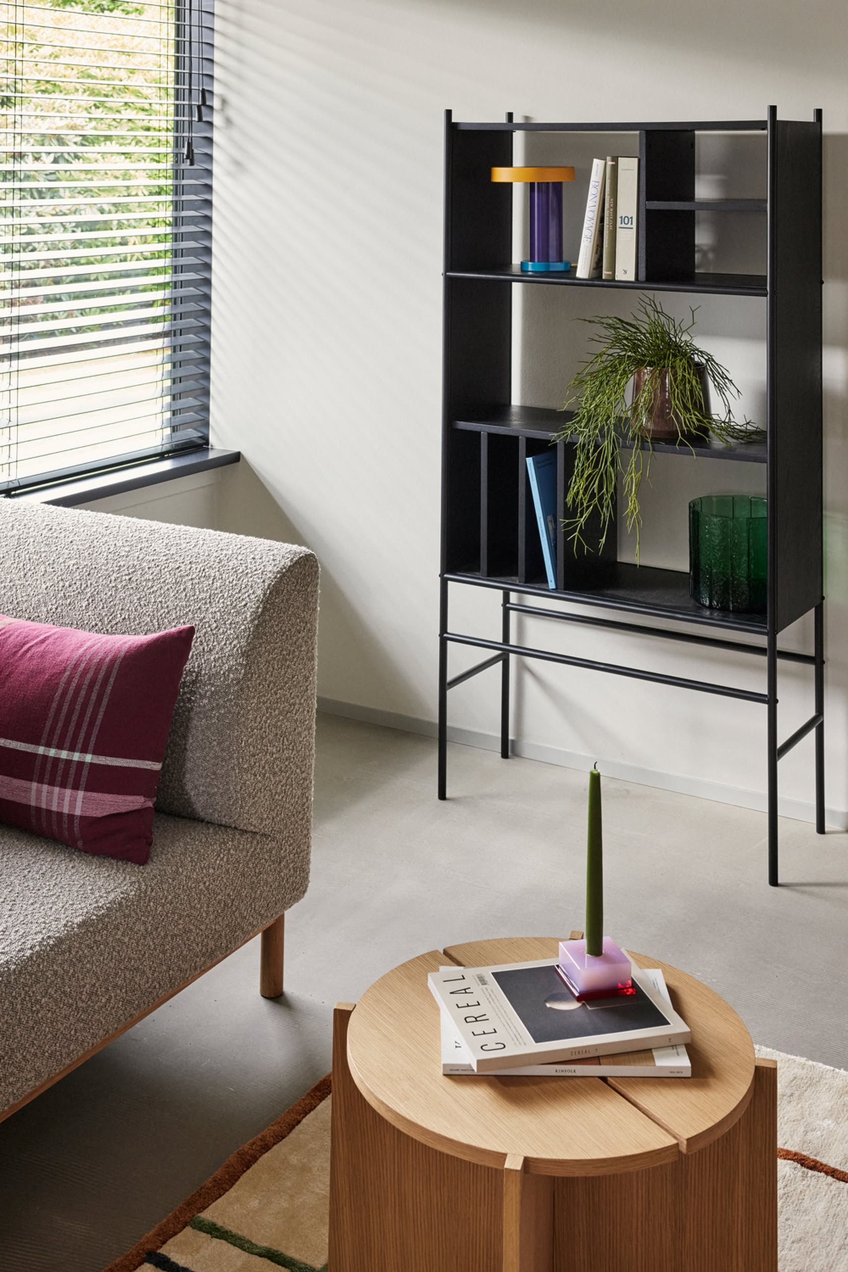 A modern living room with a textured gray sofa, a minimalist Hübsch Dash Side Table by Hübsch made from FSC-certified wood displaying books and a green candle, a black shelving unit with books and a plant, and sunlight filtering through window blinds.