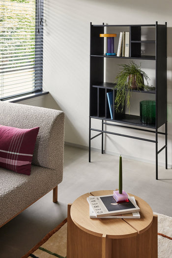 A modern living room with a textured gray sofa, a minimalist Hübsch Dash Side Table by Hübsch made from FSC-certified wood displaying books and a green candle, a black shelving unit with books and a plant, and sunlight filtering through window blinds.