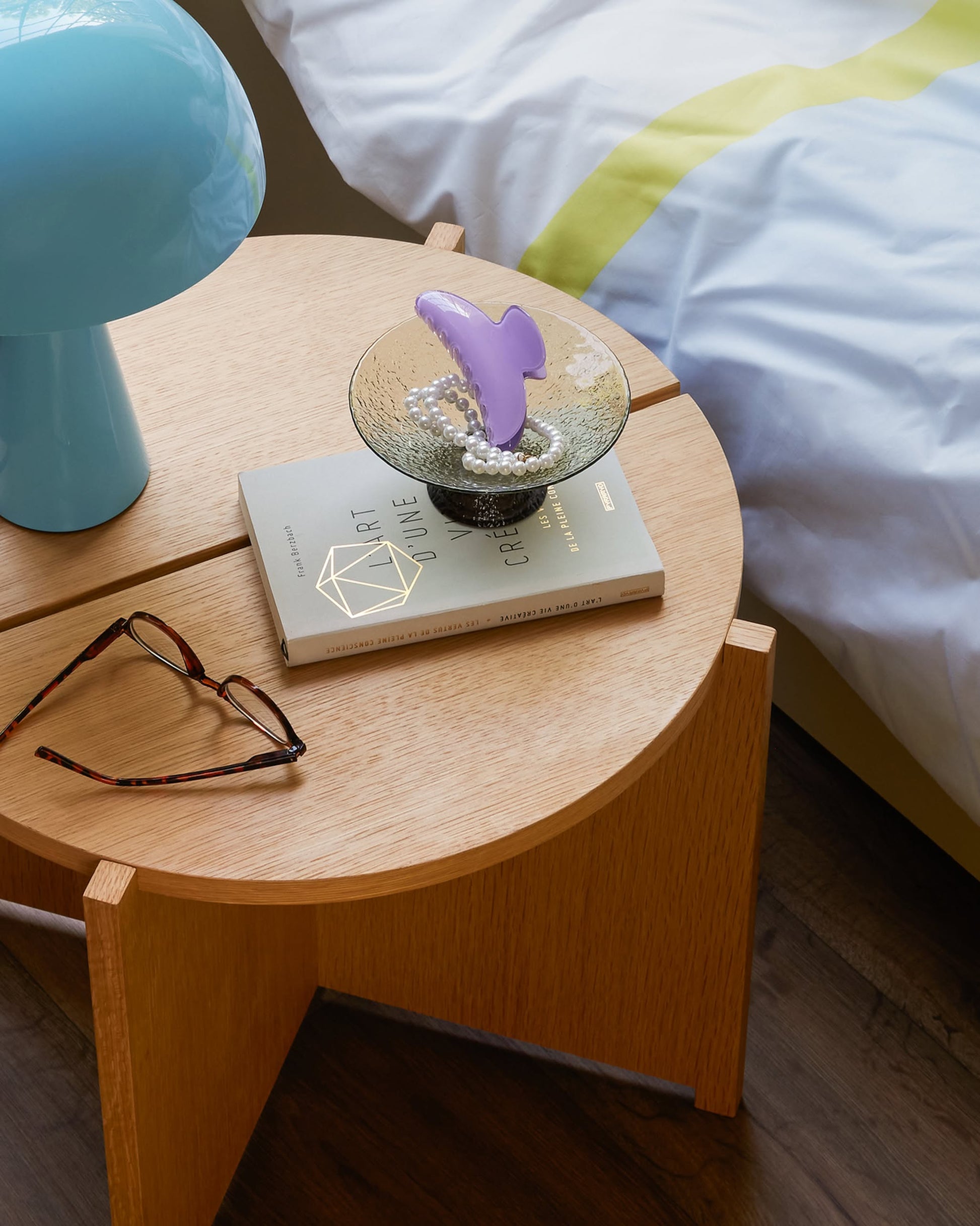 A round Hübsch Dash Side Table made of FSC-certified wood holds a blue lamp, eyeglasses, a book, and a decorative bowl with pearls and a purple object. Part of a bed with white and yellow bedding is visible nearby.