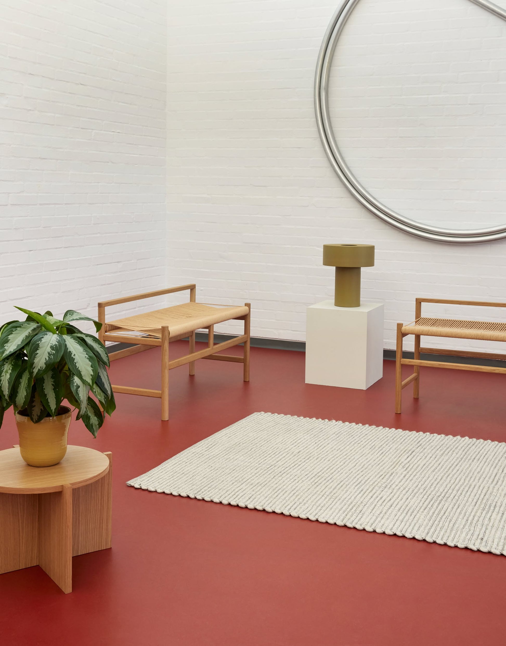 Minimalist room with red floor, white brick walls, two FSC-certified wooden chairs, a Hübsch Dash Side Table with a potted plant, modern lamp on white pedestal, cream rug, and large circular wall sculpture.