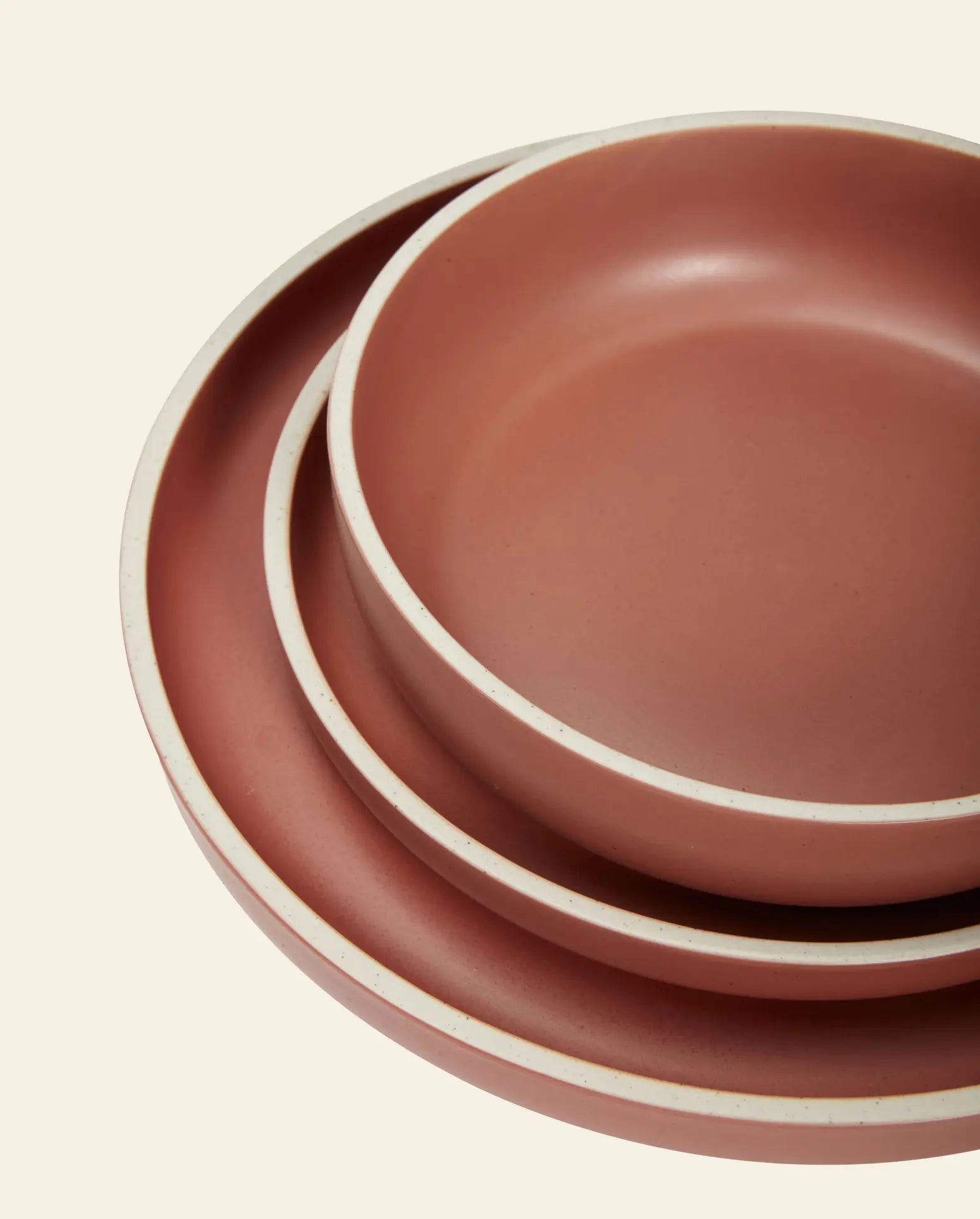 The Hübsch Dawn Bowls, Set of 3 by Hübsch, features rose-hued ceramic bowls with white rims stacked overhead on a beige background, showcasing minimalist elegance.