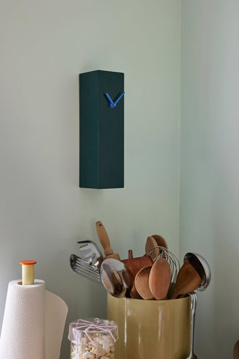 The Hübsch Hour Clock by Hübsch, featuring blue hands and a unique rectangular design, hangs above the kitchen counter where utensils, a paper towel roll, and a jar of marshmallows are neatly arranged.