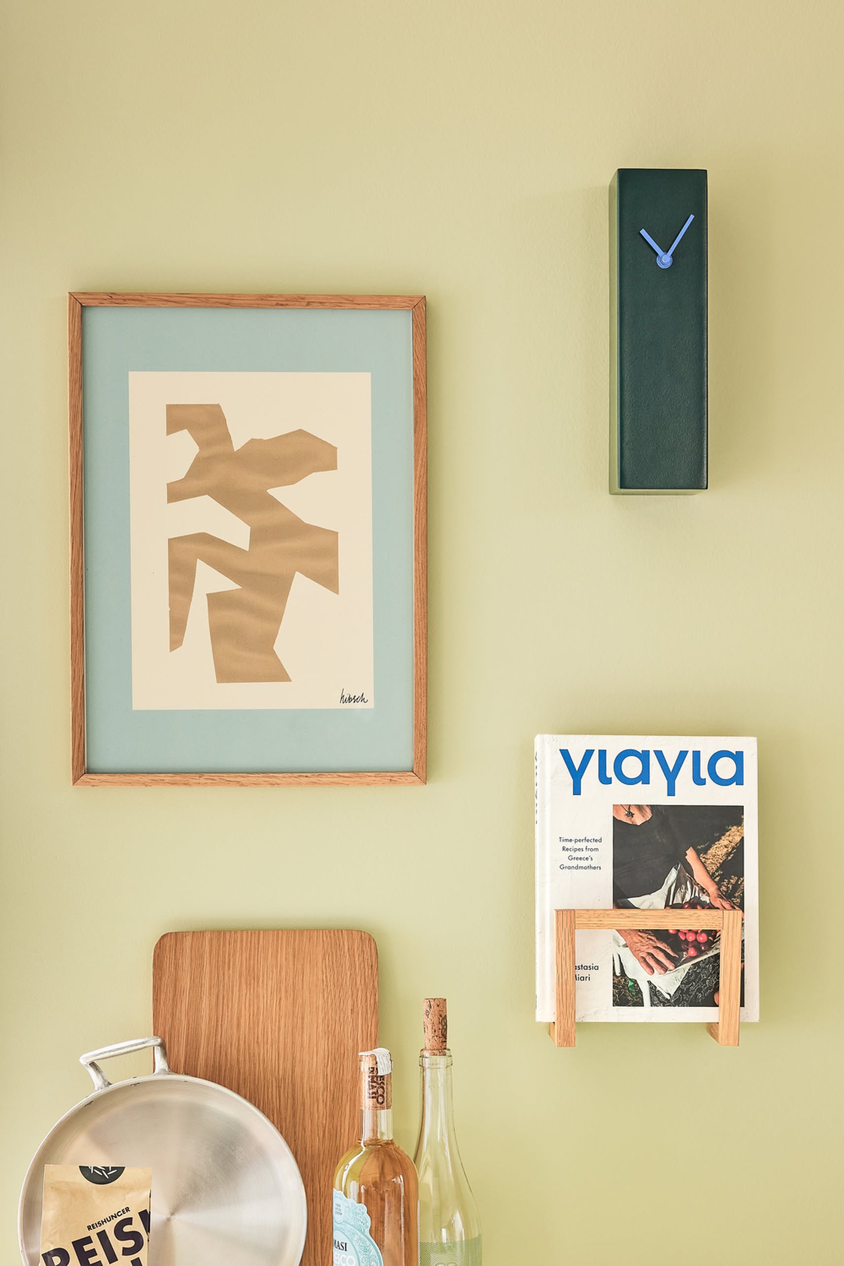 A minimalist wall displays abstract art in a wooden frame, the Hübsch Hour Clock by Hübsch with its unique modern design, a magazine on a rack, bottles, and kitchen items set against a light green background.