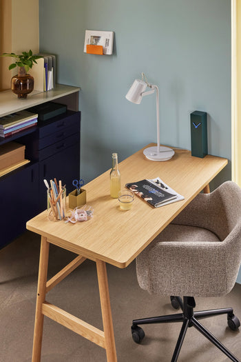 A tidy home office features a wooden desk with stationery, scissors, a drink bottle, glass, magazine, and white lamp. A cozy gray chair sits by the desk; shelves with books and the Hübsch Hour Clock by Hübsch are in the background.