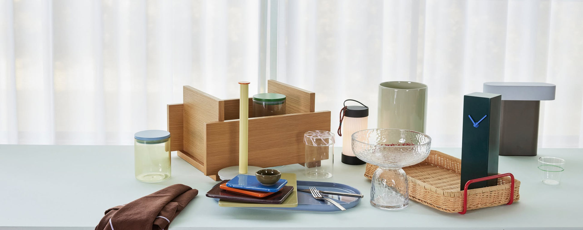 A selection of modern tableware, decor, the Hübsch Hour Clock by Hübsch, jars, a rattan basket, vases, and a wooden organizer are arranged on a light blue table set before sheer white curtains.