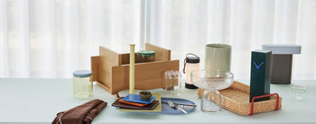 A selection of modern tableware, decor, the Hübsch Hour Clock by Hübsch, jars, a rattan basket, vases, and a wooden organizer are arranged on a light blue table set before sheer white curtains.