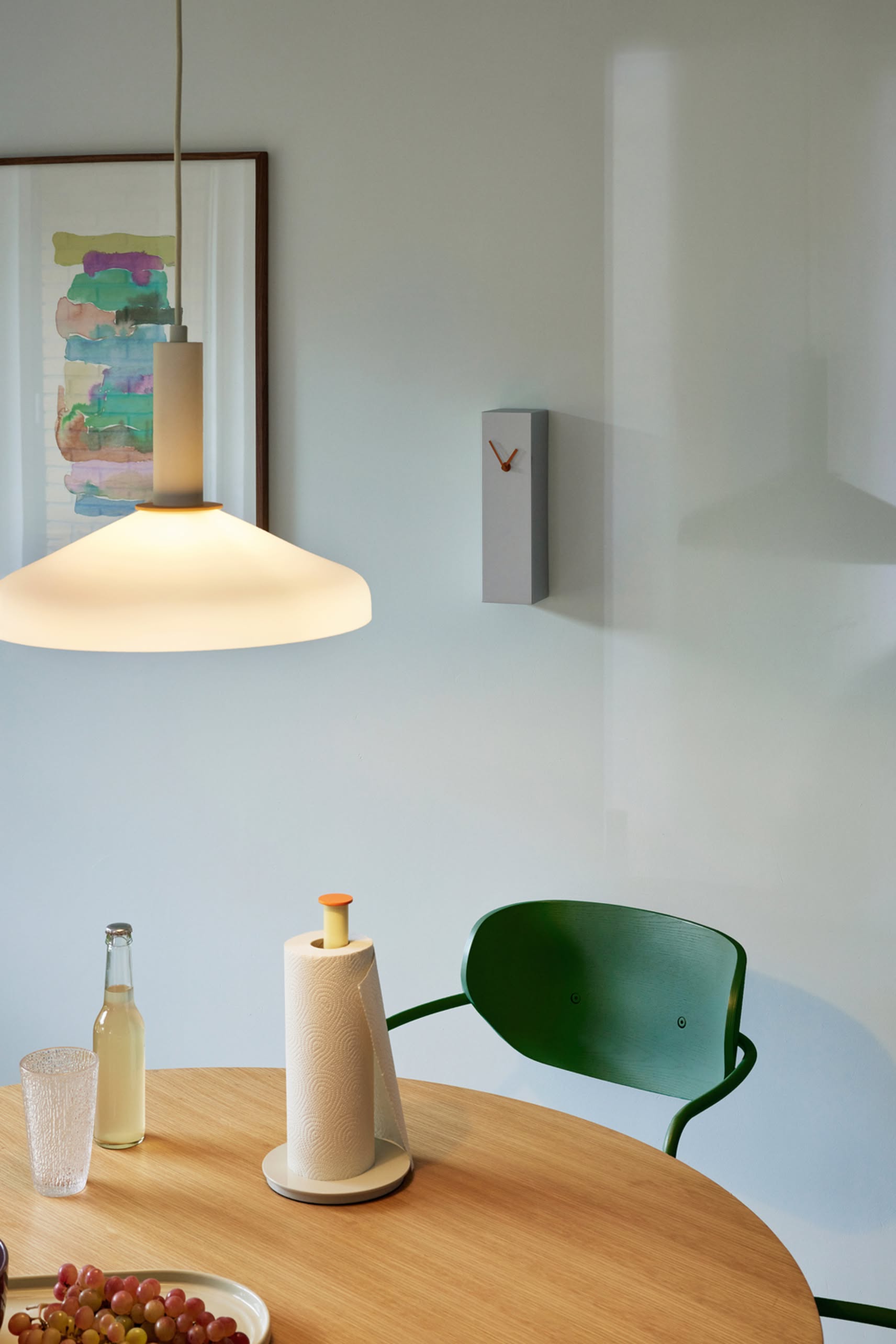 A modern dining area with a round wooden table, green chair, pendant lamp, paper towels, a glass, fruit, abstract wall art, and the minimalist Hübsch Hour Clock by Hübsch featuring a unique clock design.