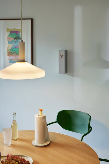 A modern dining area with a round wooden table, green chair, pendant lamp, paper towels, a glass, fruit, abstract wall art, and the minimalist Hübsch Hour Clock by Hübsch featuring a unique clock design.