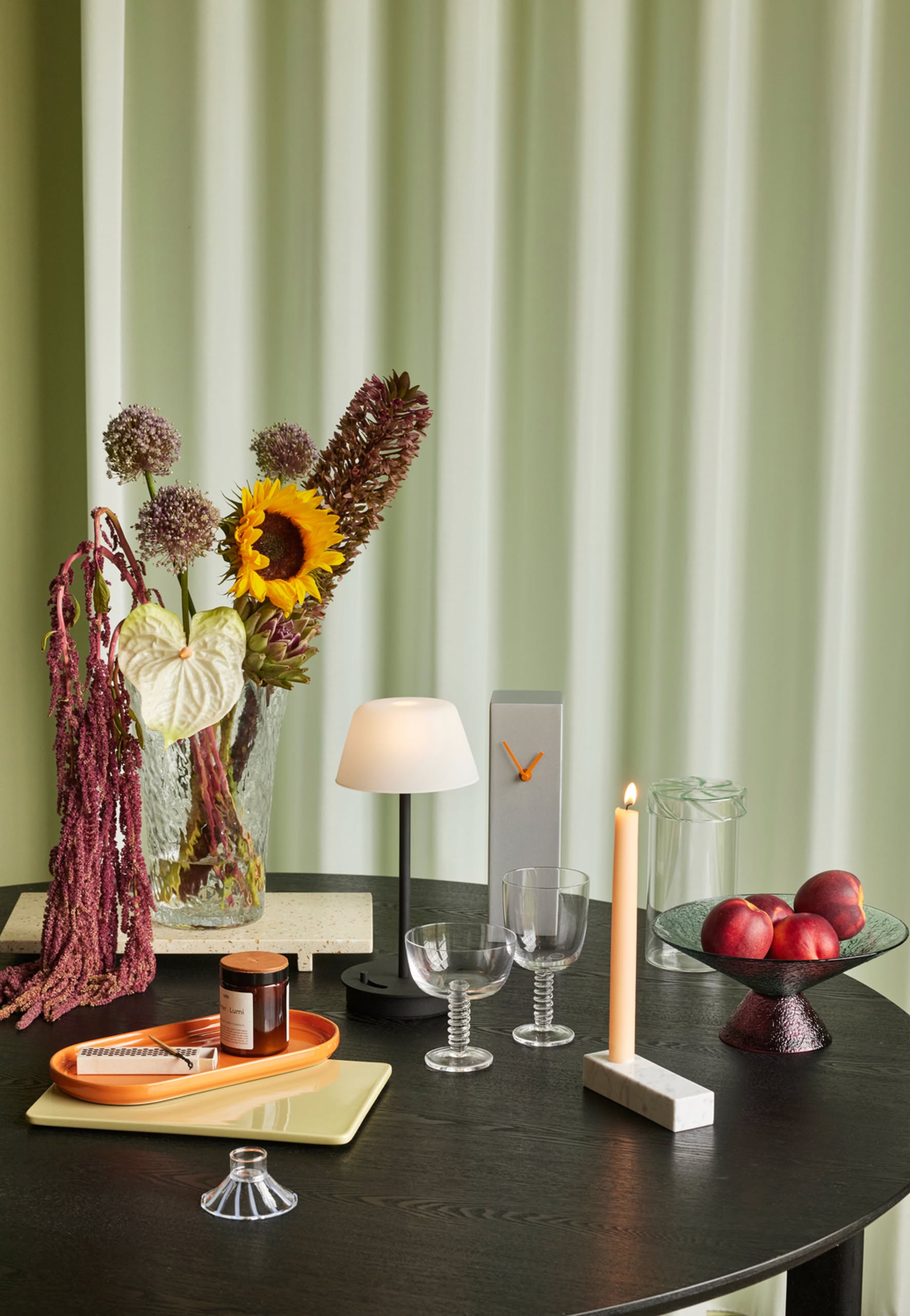 A black table holds a lamp, two ribbed wine glasses, a candle, nectarines in a bowl, a vase with sunflowers and hanging amaranth, trays with small jars, and the Hübsch Hour Clock by Hübsch, all set before pale green curtains.