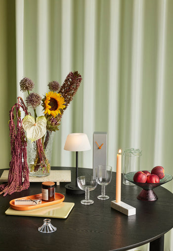 A black table holds a lamp, two ribbed wine glasses, a candle, nectarines in a bowl, a vase with sunflowers and hanging amaranth, trays with small jars, and the Hübsch Hour Clock by Hübsch, all set before pale green curtains.