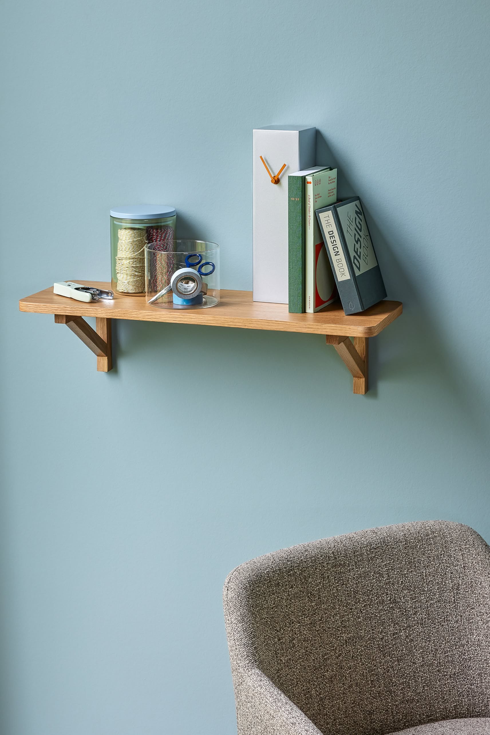 A wooden wall shelf holds books and supplies, with the Hübsch Hour Clock by Hübsch mounted above a gray upholstered chair against a light blue wall.