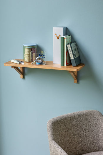 A wooden wall shelf holds books and supplies, with the Hübsch Hour Clock by Hübsch mounted above a gray upholstered chair against a light blue wall.