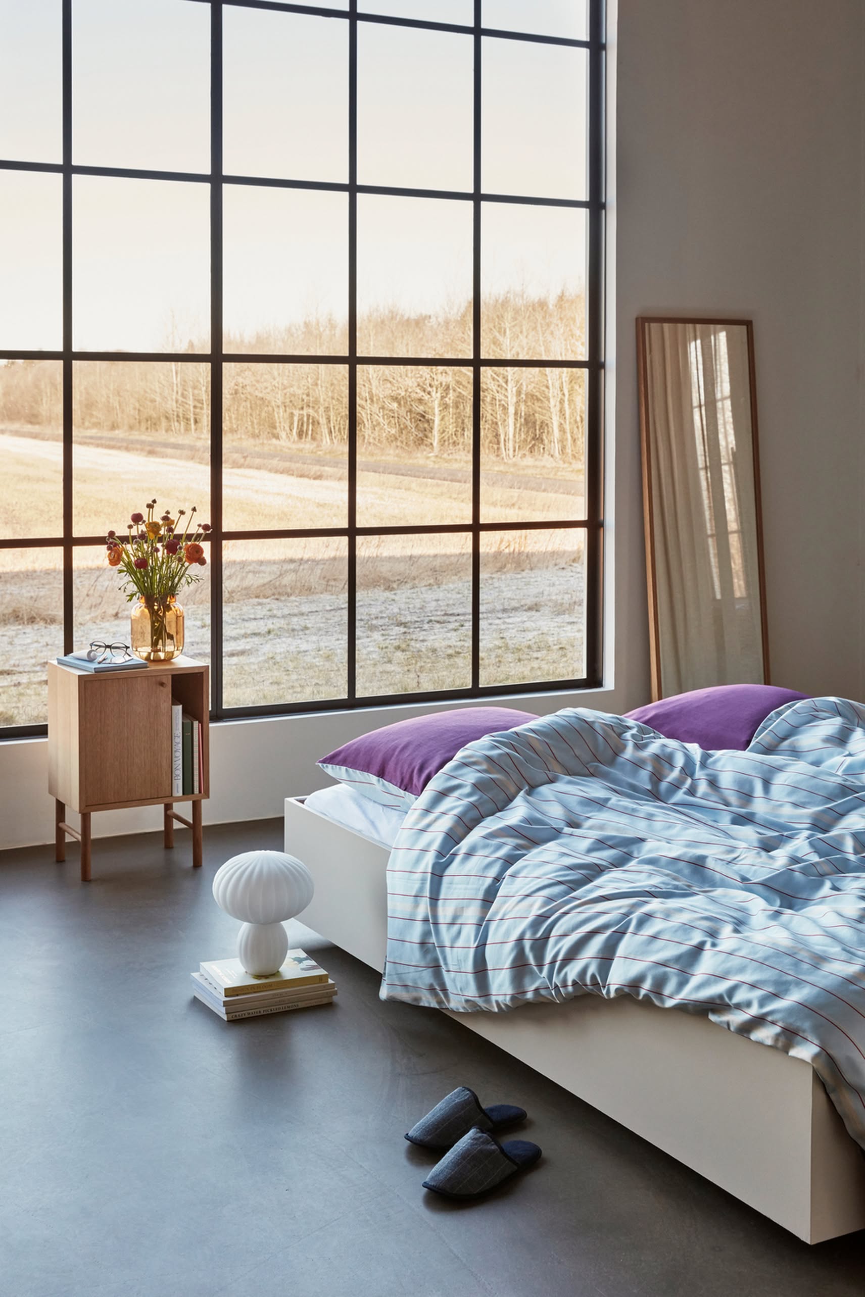 A modern bedroom with a large window overlooking a field, striped bedding, purple pillows, a wooden nightstand with flowers, the Hübsch Kumu Table Lamp by Hübsch on books, a mirror, and dark slippers for a modern minimalistic look.
