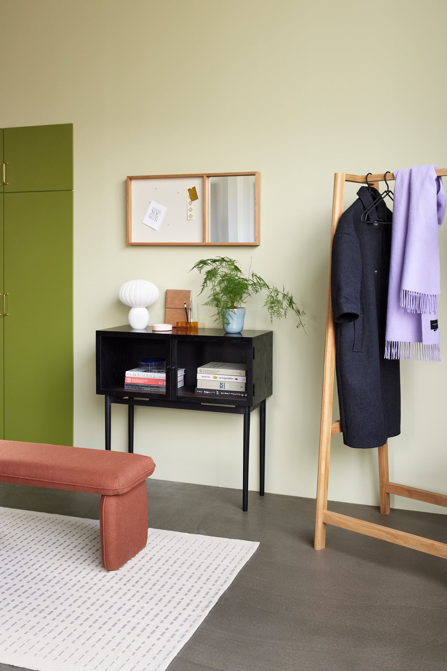 A modern minimalistic entryway features a green cabinet, wall mirror, black console with books and the Hübsch Kumu Table Lamp with dimmable function, potted plant, wooden coat rack with dark coat and lavender scarf, rust bench, and white rug on gray floor.