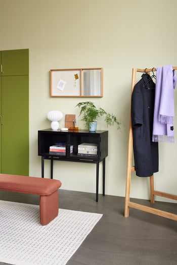 A modern minimalistic entryway features a green cabinet, wall mirror, black console with books and the Hübsch Kumu Table Lamp with dimmable function, potted plant, wooden coat rack with dark coat and lavender scarf, rust bench, and white rug on gray floor.