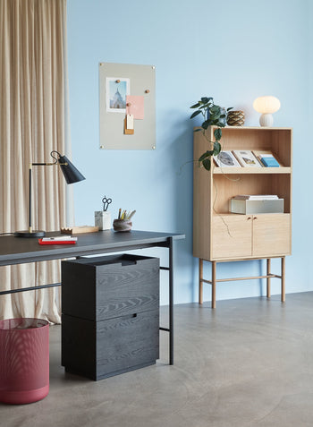 A modern home office with a black desk, organizer, and the Hübsch Kumu Table Lamp by Hübsch featuring dimmable light, set against a light blue wall. A wooden bookshelf holds books and a plant. Beige curtains complete the cozy, minimalistic look.