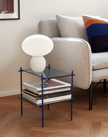 A modern living room corner features a light gray armchair, colorful pillow, and a glass side table displaying the Hübsch Kumu Table Lamp by Hübsch with dimmable function, alongside stacked magazines on a wooden floor.