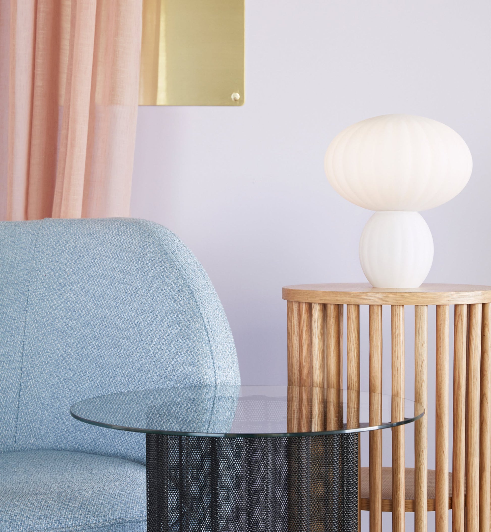 A modern minimalistic look features a light blue upholstered chair, round glass coffee table, wooden side table with the Hübsch Kumu Table Lamp by Hübsch, pale pink curtains, and a gold wall accent on a light-colored wall.