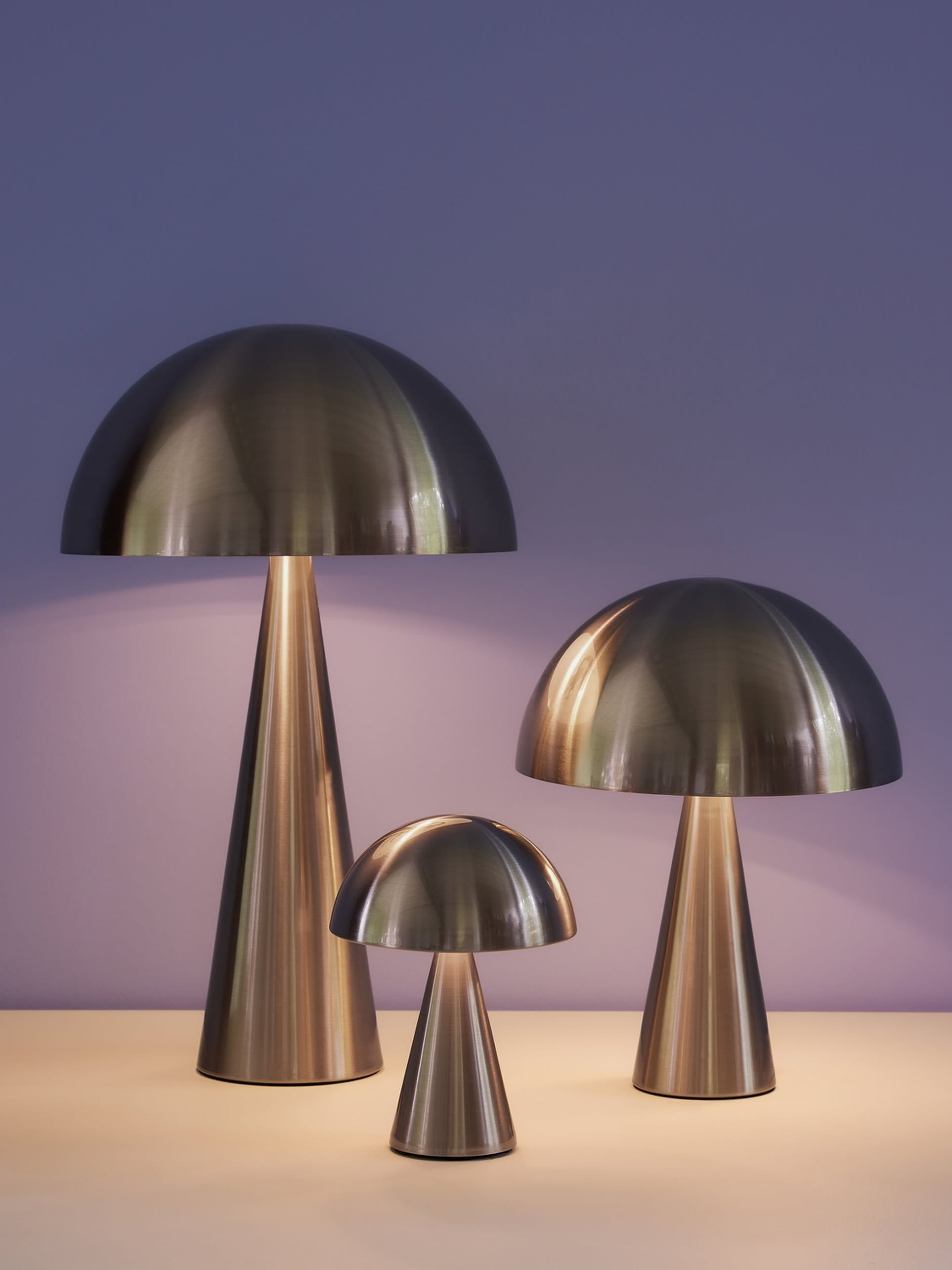 Three Hübsch Mush Table Lamp Mini lights of varying heights are displayed on a light surface with a soft purple background. Featuring conical bases and warm glow, these modern lamps by Hübsch come in multiple color options.