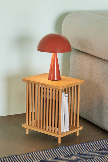A small wooden side table with vertical slats holds the Hübsch Mush Table Lamp Mini by Hübsch, adding a pop of color to this modern home. Magazines fill the table, which sits by a beige sofa on a blue and beige striped rug.