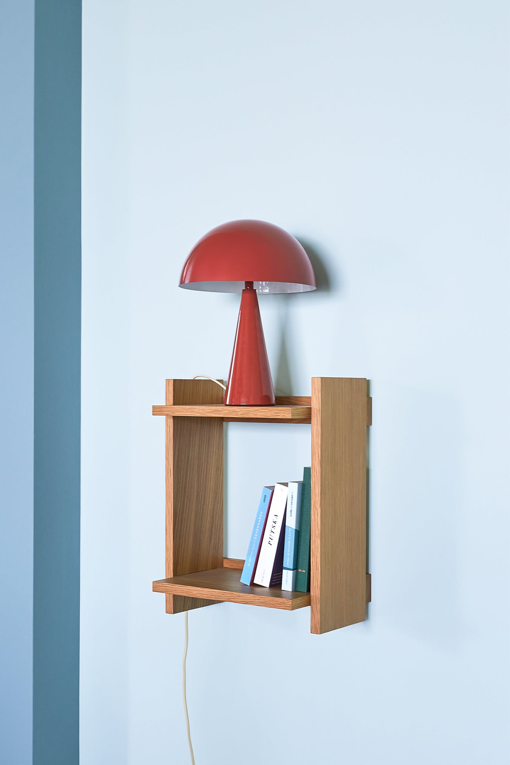A Hübsch Mush Table Lamp Mini in red by Hübsch sits on a small wooden wall shelf above stacked books, adding a pop of color and modern flair to the light blue wall.
