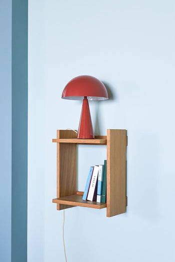 A Hübsch Mush Table Lamp Mini in red by Hübsch sits on a small wooden wall shelf above stacked books, adding a pop of color and modern flair to the light blue wall.